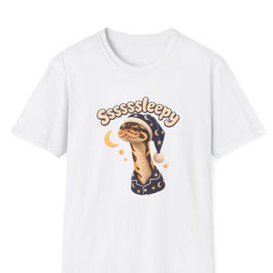 May include: A white short-sleeved t-shirt featuring a graphic of a snake wearing a dark blue nightcap and pajamas with a moon and star pattern. The text Sssssleepy is printed in a light brown font above the snake, accompanied by a crescent moon.