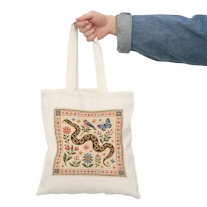 May include: A cream-colored canvas tote bag featuring a central square graphic print. The design displays a winding brown and black snake surrounded by a blue bird, a blue butterfly, pink flowers, and small star motifs, all framed by a geometric border.