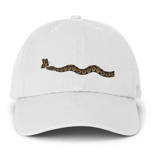 May include: A white baseball cap featuring a graphic print of a brown and black patterned snake moving in a wavy line towards a small yellow and black butterfly positioned near the snake's head on the front centre of the crown.
