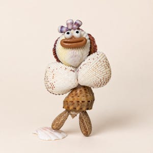 May include: A decorative figurine crafted from various seashells, featuring a head made of a white shell with a brown mouth and two round eyes, a torso of two large white shells, and a lower body of a brown patterned shell with two elongated legs.