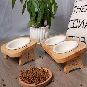 Peut inclure: Deux stations de nourriture pour animaux en bois clair avec des bols en céramique blanche. Une station contient un bol, l'autre deux. Un petit plat en bois rempli de croquettes brunes est posé devant, avec une empreinte de patte gravée.