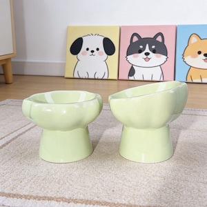 May include: Two light green ceramic pet bowls with a raised, pedestal-style base and a scalloped, paw-shaped rim. The bowls feature a smooth, glossy finish and are designed with an angled opening to provide an elevated feeding position.