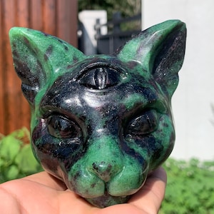 May include: A carved stone cat head sculpture featuring a third eye on the forehead. The material is a mottled green and black mineral with a polished, smooth surface. The feline features include pointed ears, almond-shaped eyes, and a nose.
