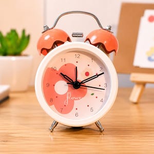 May include: A round analog alarm clock with a white frame and two orange bells on top. The clock face features a light pink background with a large coral-colored flower graphic, black hour and minute hands, and the word flower written in script.
