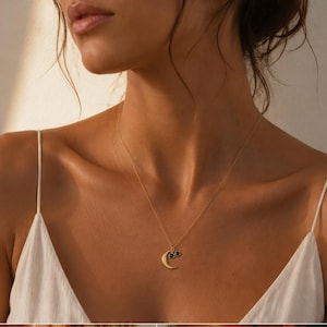 May include: A gold-toned necklace featuring a delicate chain with a crescent moon pendant and two small, circular charms, one set with a dark green stone and the other with a dark blue stone, resting against a white fabric garment.