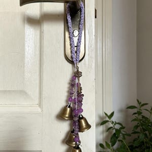 Peut inclure: Une sangle crochetée lavande suspendue à une poignée de porte, avec une breloque clé couleur bronze et plusieurs brins de pierres précieuses violettes alternés avec quatre petites clochettes en laiton poli de longueurs variées.