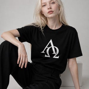 Może przedstawiać: Czarny t-shirt z krótkim rękawem z dużym białym nadrukiem graficznym na klatce piersiowej. Wzór składa się ze stylizowanej wielkiej litery A nałożonej na grecką literę Omega. Koszulka posiada okrągły dekolt oraz standardowy krój.