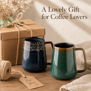 May include: Two ceramic mugs with handles, one dark blue with white vertical dashes and one green with a brown rim. A brown gift box with twine, a tag reading "For You" with a heart, and text "A Lovely Gift for Coffee Lovers" are visible.