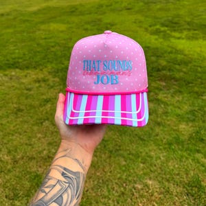 May include: A pink trucker hat featuring a white polka dot pattern on the crown and a vibrant vertical striped brim in shades of pink, purple, and light blue. The text "THAT SOUNDS like a man's JOB" is embroidered in blue and pink on the front.