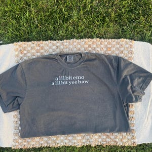 May include: A charcoal gray short-sleeve t-shirt featuring white text printed on the chest that reads a lil bit emo a lil bit yee haw. The shirt has a crew neckline and a standard fit, showcasing a simple design with a clear, legible font.