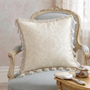 May include: A square cream-colored throw pillow featuring an intricate tone-on-tone damask pattern. The edges are finished with a decorative braided trim and small hanging tassels along the perimeter, creating a textured and detailed appearance.