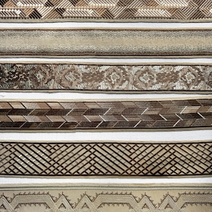 May include: Seven horizontal strips of decorative fabric trim arranged in a stack. The designs include a Greek key pattern, a woven geometric texture, a metallic shimmer band, a floral damask, a chevron weave, a lattice grid, and a diamond motif.