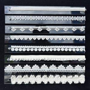 May include: Eight distinct strips of white lace trim arranged horizontally. Each strip features a unique pattern, including floral, scalloped, and geometric designs. The lace pieces are mounted on clear rectangular supports against a black background.