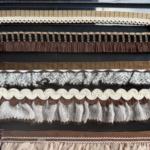 May include: A collection of decorative trim ribbons arranged in horizontal rows. The assortment includes a white pom-pom trim, a tan plaid ribbon, a brown fringe trim, a textured gray ruffled ribbon, a cream gimp trim, and a beige fabric fringe.