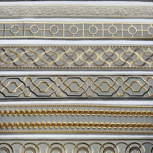Peut inclure: Une collection de huit rubans décoratifs en tissu disposés horizontalement. Chaque ruban présente des broderies complexes dans des tons dorés, bronze et beige, illustrant divers motifs géométriques, volutes et lignes répétitives.
