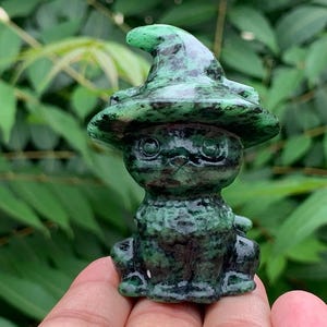 May include: A small, polished green and black stone carving of a cat wearing a pointed witch hat. The figurine features a smooth, glossy texture with dark mottled patterns throughout the mineral surface, depicting a seated cat with large eyes.