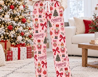 May include: Light pink pajama pants featuring a repeating Christmas pattern of nutcrackers, decorated pine trees, red bows, and ornaments. The name Charlotte is printed in red script throughout the fabric. The pants have a white drawstring waist.