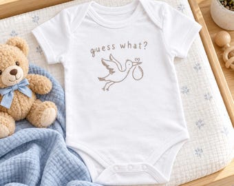 May include: A white short-sleeved baby bodysuit featuring a brown line-art illustration of a stork carrying a bundle and the text "guess what?" printed in brown. The garment has a round neckline and three snap closures at the bottom crotch area.