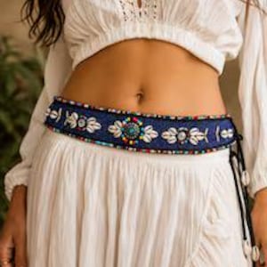 May include: A dark blue fabric belt featuring a central row of white cowrie shells, small turquoise-coloured beads, and multi-coloured seed beads. The belt is secured with thin black side ties that are finished with dangling white cowrie shell accents.