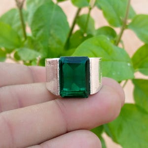 May include: A silver-toned ring featuring a large, rectangular, emerald-green faceted gemstone set in the center. The band is wide and smooth with a metallic finish, providing a bold contrast to the vibrant, deep green color of the central stone.