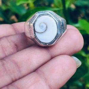 Puede incluir: Un anillo de tono plateado con una incrustación ovalada de concha de ojo de shiva blanca. El engaste de metal tiene un marco octogonal con detalles texturizados y granulados en los bordes, rodeando el patrón en espiral liso de la concha.