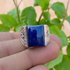 May include: A silver ring featuring a rectangular, deep blue lapis lazuli gemstone set in the center. The band is crafted with intricate, raised metalwork patterns on both sides, showcasing a detailed, textured design along the shank.