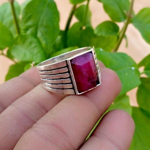 May include: A silver ring featuring a rectangular, faceted deep red gemstone set in the center. The band of the ring is constructed with four horizontal parallel lines that wrap around the sides, creating a layered, metallic appearance.