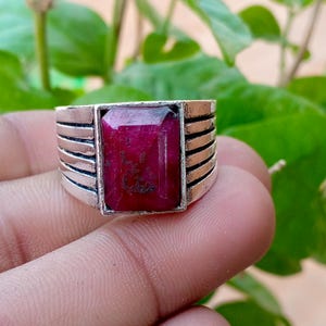 May include: A silver-toned ring featuring a rectangular, deep red gemstone set in the centre. The band is constructed with five horizontal, parallel metal bars on each side of the stone, creating a layered, ribbed appearance around the finger.
