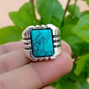 Puede incluir: Un anillo de tono plateado con una piedra turquesa rectangular brillante con vetas oscuras e intrincadas. La banda de metal tiene un diseño texturizado y en capas a ambos lados de la piedra central, creando una apariencia geométrica audaz.