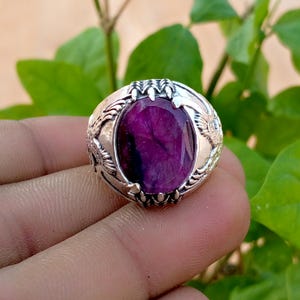 May include: A silver ring featuring a large, oval-shaped, deep magenta gemstone set within a metal band. The setting is designed with intricate, claw-like prongs that grip the stone, and the band displays detailed, textured metalwork on the sides.