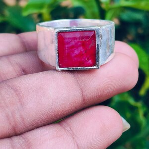 May include: A silver ring featuring a square-cut, deep red gemstone set in a smooth, polished metal band. The setting is rectangular and holds the vibrant red stone securely in the centre, showcasing a clean, metallic finish on the wide band.