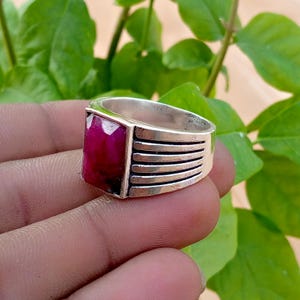 May include: A silver ring featuring a rectangular, faceted, deep pink gemstone set in the center. The band of the ring is constructed with a series of five horizontal, parallel grooves that wrap around the sides of the metallic silver shank.