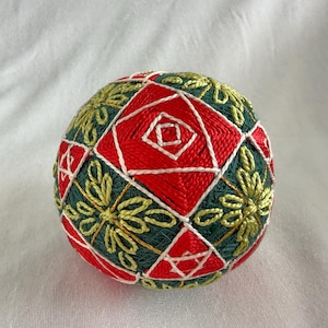 May include: A spherical temari ball featuring intricate embroidery in vibrant red, deep forest green, and bright yellow thread. The surface displays a geometric pattern of squares and triangles outlined in white, accented by floral-like motifs.