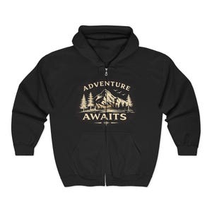 May include: A black zip-up hoodie featuring a cream-colored graphic on the chest. The design depicts a mountain range, pine trees, and birds, with the text ADVENTURE AWAITS printed in a bold, capitalized font above and below the illustration.