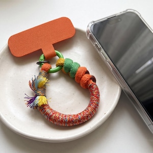 May include: A phone tether tab in bright orange attached to a green plastic ring. The ring is wrapped in multicolored cord with sections of solid green, orange, and brown, featuring a frayed tassel end with yellow, blue, and purple threads.