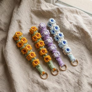 May include: Four macrame wristlets featuring a repeating floral pattern. Each wristlet is crafted from knotted cord in distinct color combinations: yellow and orange, orange and yellow, purple and white, and light blue and white, each with a gold ring.