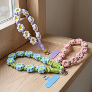 May include: Three macrame wrist straps featuring daisy-shaped knots in lavender, light blue, and pink. Each strap includes a silver metal ring attached to a flat, solid-colored plastic tab in matching lavender, light blue, or pink tones.
