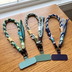 May include: Three braided cord wrist straps with tassels, each attached to a black metal lobster clasp and a rectangular flat connector piece. The straps feature varied color palettes of green, yellow, blue, and purple threads in a woven design.