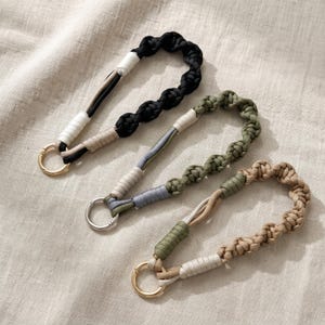 May include: Three wristlet keychains made of braided cord. The top one is black with white and tan accents and a gold ring. The middle one is olive green with grey and cream accents and a silver ring. The bottom one is tan with olive and cream accents and a gold ring.