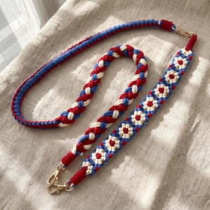 May include: A macrame strap featuring a braided design in red, white, and light blue cord. One section displays a repeating floral pattern, while the other is a simple braid. The strap includes gold-toned metal swivel clasps at both ends.