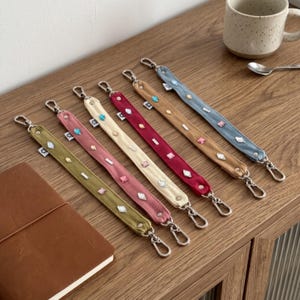 May include: Six fabric wristlet straps in olive green, dusty pink, cream, deep red, tan, and slate blue. Each strap features silver-tone metal lobster clasps at both ends and is decorated with various small, colorful, geometric-shaped rhinestones.
