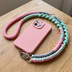 May include: A light pink smartphone case with a triple-lens camera cutout, attached to a long, braided cord strap. The strap features a mix of pastel pink, mint green, and metallic silver cord sections, secured with silver-toned metal lobster clasps.