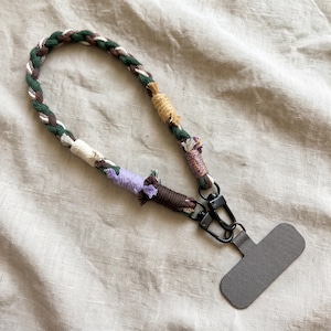 May include: A braided phone lanyard featuring a mix of green, brown, white, yellow, and lavender cotton cords. The strap is accented with wrapped thread details and connects to a gray rectangular phone tether tab via two black metal lobster clasps.