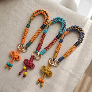 May include: Three braided paracord wrist lanyards featuring gold-tone metal rings and decorative knot ends. The first is orange and dark green, the second is teal, maroon, and yellow, and the third is navy blue, mustard yellow, and orange.