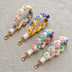 May include: Four macrame wristlet keychains featuring floral patterns made from knotted cord. Each keychain includes a gold-toned metal lobster clasp. The designs showcase various color combinations including beige, yellow, blue, pink, and pastel tones.