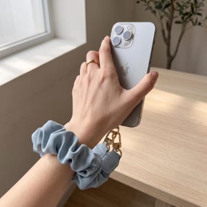 Könnte beinhalten: Ein hellblaues Stoff-Handgelenkband im Scrunchie-Stil mit goldfarbenen Metallclips, befestigt an einem silbernen Smartphone. Das Armband weist eine geraffte, elastische Textur auf und wird um das Handgelenk getragen.