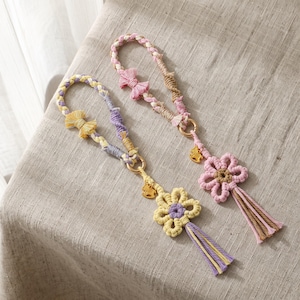 May include: Two macrame wristlet keychains featuring braided loops with small fabric bows. One keychain has yellow and purple accents with a flower charm, while the other has pink and tan accents with a pink flower and a gold-toned charm.