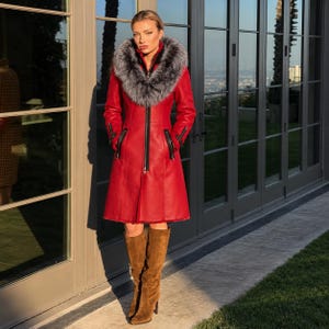May include: A vibrant red knee-length coat featuring a large, fluffy grey and white faux fur collar, a central black zipper closure, and black zippered pockets on the sides. The coat is paired with knee-high brown suede boots with a block heel.
