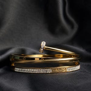 Könnte beinhalten: Ein goldfarbenes Metallarmband in Nagelform mit einem mit klaren Steinen besetzten Kopf und einem teilweise mit klaren Steinen ausgefassten Band. Das Wort Cartier ist an der Seite des Bandes eingraviert, das eine polierte Oberfläche aufweist.