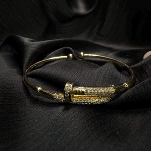 May include: A gold-toned metal bangle bracelet shaped like a nail, featuring a curved band that wraps around the wrist. The head and the pointed end of the nail design are encrusted with small, clear, sparkling stones along the top surface.